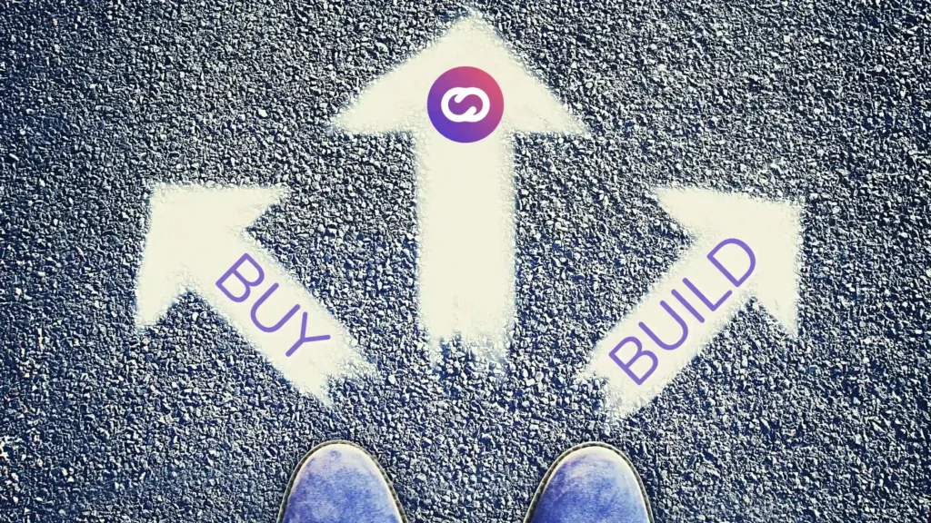 A top-down view of three white arrows painted on asphalt, pointing in different directions with the words “BUY” and “BUILD” on the side arrows, and a central arrow marked with the Cloud Studio logo. A pair of feet stands at the decision point, symbolizing a choice between building or buying an IoT solution.