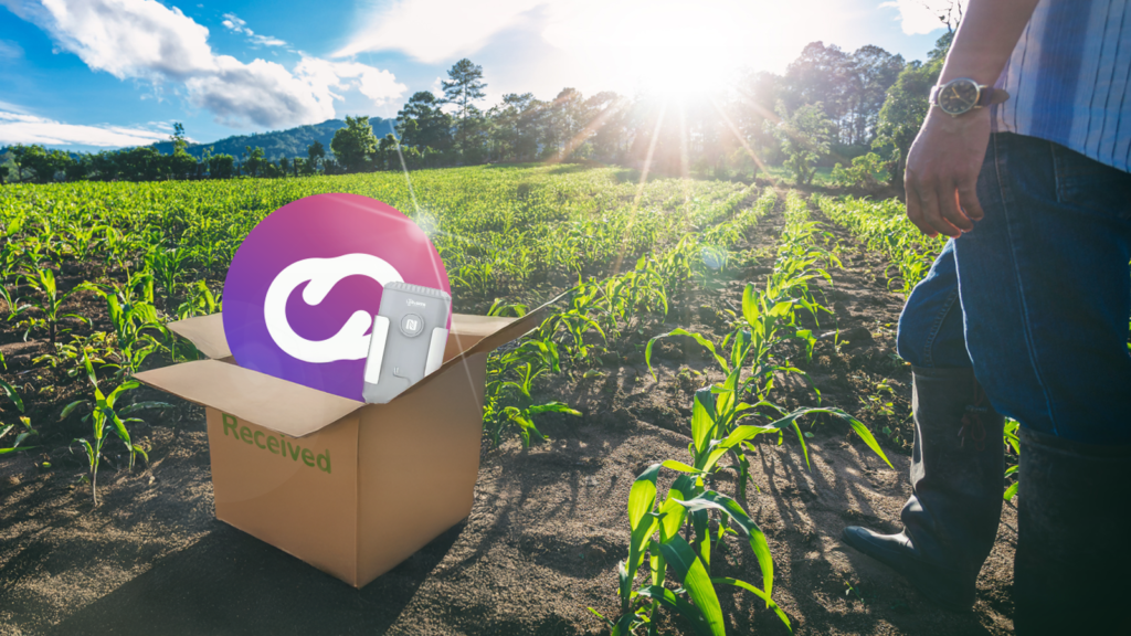 Una caja abierta con el logo de Cloud Studio y un sensor IoT en medio de un campo de cultivo soleado, simbolizando tecnología de riego inteligente lista para optimizar el uso de agua en agricultura.